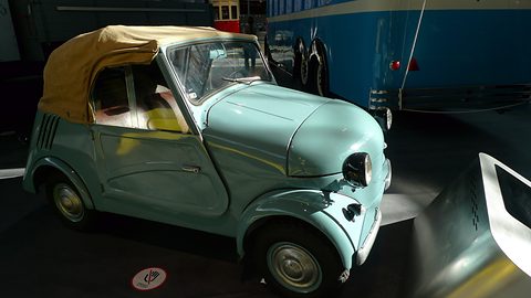 Low view of a pale blue microcar with its beige hood up, a headlamp on its front wing, a blue tram behind. Opens a larger view