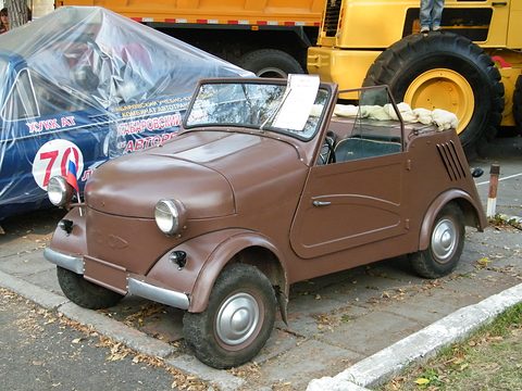 Brown open microcar on a kerbside parking strip, a covered rally car and a big yellow construction machine behind. Opens a larger view
