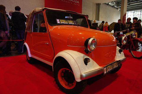 Orange microcar with cream mudguards and a beige hood on a red carpet, a red motorcycle and visitors beside it. Opens a larger view