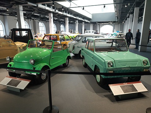 A bright green open microcar and a pale green boxy two-door microcar side by side behind a rope, more small cars beyond. Opens a larger view