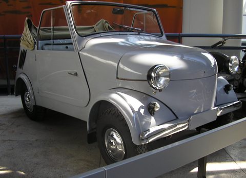Pale grey open microcar with a folded hood seen from the front right, museum barriers in front. Opens a larger view