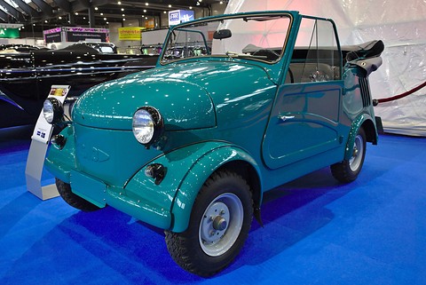 Glossy teal open microcar with a headlamp on each front wing and a folded black hood, on a blue show carpet. Opens a larger view