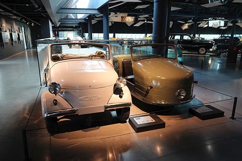 A pale pink open four-wheeled microcar and a tan bulbous three-wheeler side by side on a lit museum plinth.