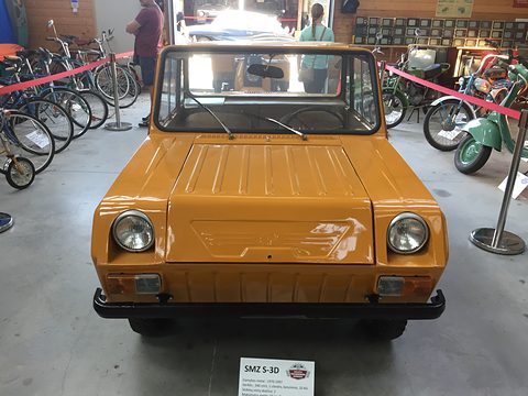 Front of an ochre boxy microcar with round headlamps and a ribbed bonnet, rows of bicycles and motorcycles either side. Opens a larger view