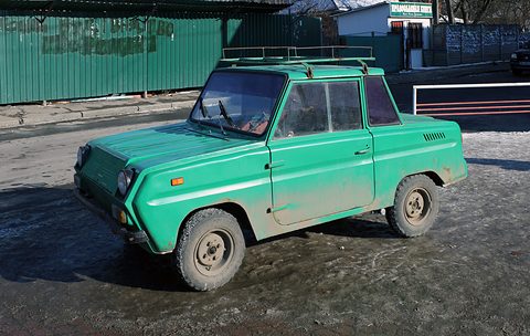 Dusty green boxy microcar with a roof rack parked on a muddy yard in front of a green corrugated fence. Opens a larger view