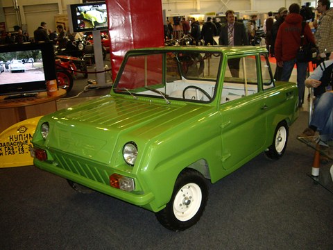 Lime-green boxy microcar with white wheels on a dark show floor, screens and visitors behind. Opens a larger view