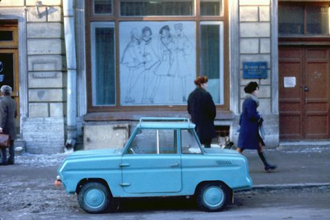 Colour photograph of a pale blue boxy microcar at a kerb, two women in winter coats walking past a shop window behind it.