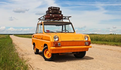 Bright orange boxy microcar on a dirt country track, two old suitcases strapped to its roof rack, open fields behind. Opens a larger view