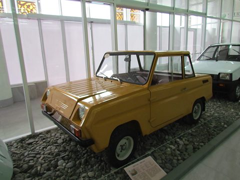 Ochre boxy microcar on a pebble bed in a glass-walled hall, a white Oka parked behind it. Opens a larger view