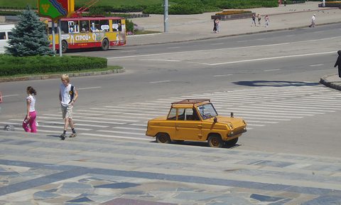 A small yellow boxy car with a roof rack crosses a zebra crossing, two people walking beside it, a trolleybus behind. Opens a larger view