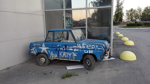 Battered blue boxy microcar daubed with white Russian slogans and a star, parked against a glass shop front. Opens a larger view