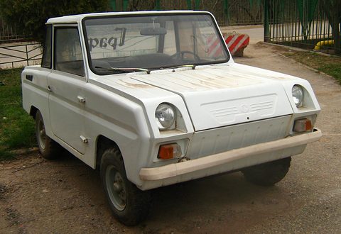 White boxy microcar with a cream bumper and round headlamps, parked on a gravel yard by a fence.