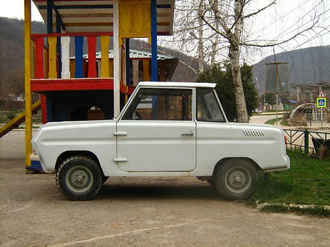 Side view of a white boxy two-door microcar on gravel, a colourful wooden play tower behind and wooded hills beyond. Opens a larger view