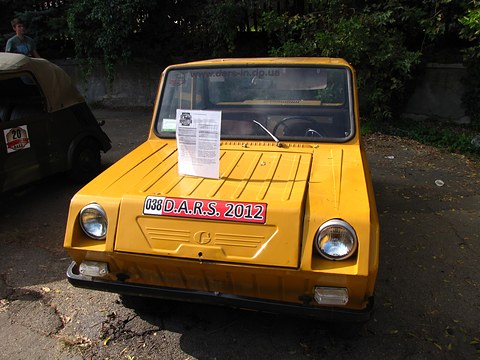Head-on view of a yellow boxy microcar in dappled shade, a rally plate on its bonnet and a paper sign in the windscreen. Opens a larger view