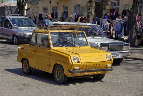 Yellow boxy microcar with a roof rack in a street, stalls of clothes and a Lada saloon behind it. Opens a larger view