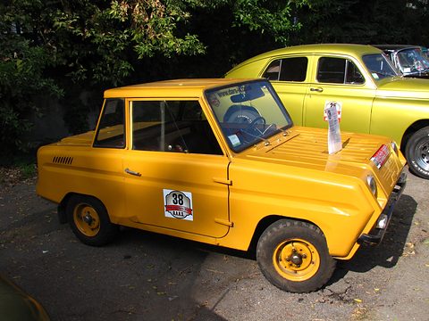 Yellow boxy microcar with a round rally sticker on its door, a lime-green saloon parked behind under trees. Opens a larger view