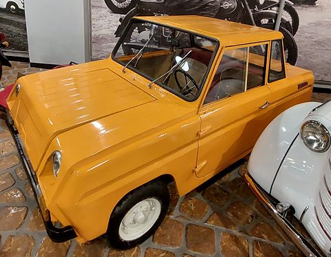 Glossy yellow boxy microcar on a cobbled museum floor, motorcycles behind and a white car’s nose beside it.