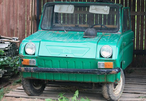 Head-on view of a worn green boxy microcar in a wooden shed, round headlamps above orange indicators, mud on its bumper.