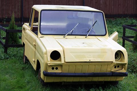 Head-on view of a faded cream boxy two-seater with empty headlamp holes and a black bumper, parked on grass by a wooden fence. Opens a larger view