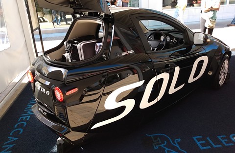 Rear of a black single-seat three-wheeler with a raised hatch showing luggage, large SOLO lettering on its side. Opens a larger view