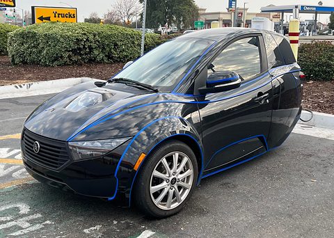 Dark blue single-seat three-wheeler with blue outline stripes parked by a hedge, a petrol station behind. Opens a larger view