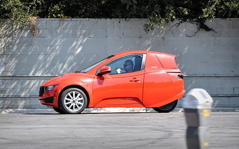 Orange single-seat three-wheeler with its driver aboard, parked by a parking meter in front of a block wall.