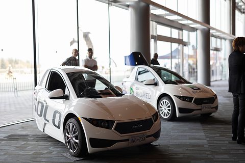 Two white single-seat three-wheelers on display in a glass-walled hall, people standing around. Opens a larger view