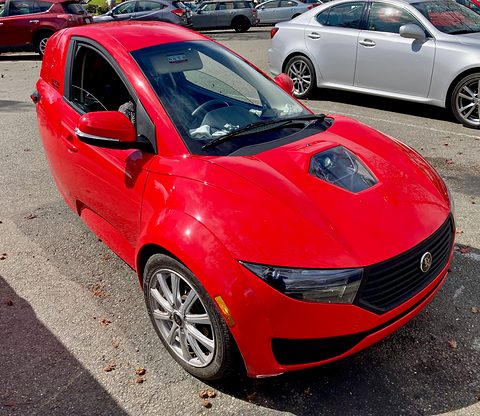 Red single-seat three-wheeler seen from above the front, with its bonnet scoop and long screen, among parked cars. Opens a larger view