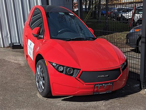 Red single-seat three-wheeler with a grey grille, parked on gravel beside a metal fence.