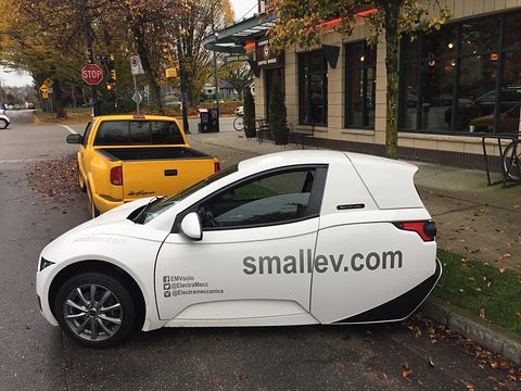 White single-seat three-wheeler with smallev.com lettering parked by a café, a yellow pick-up behind.