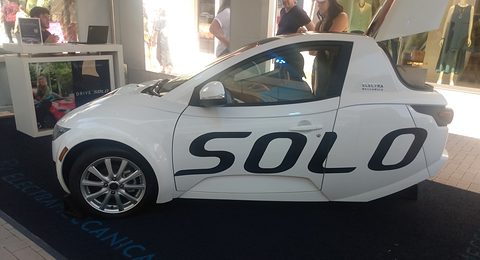 Side view of a white single-seat three-wheeler with black SOLO lettering on a dark carpet in a showroom. Opens a larger view