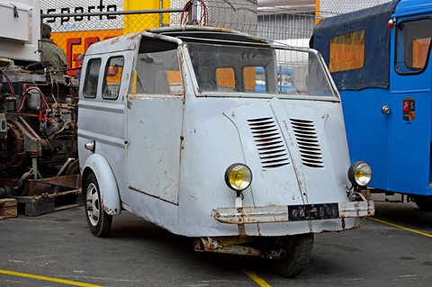 Weathered pale grey three-wheeled van with louvred nose, two round lamps and rusty bumper, a blue van beside it. Opens a larger view