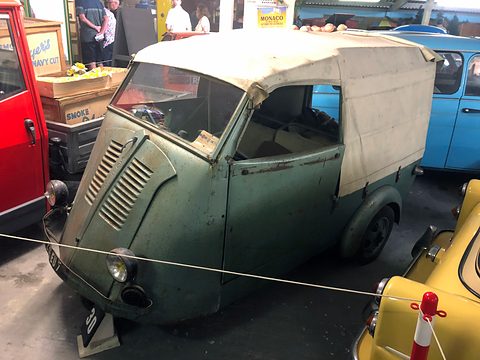 Weathered grey-green three-wheeled van with a stained cream canvas back, roped off between a red car and a yellow car. Opens a larger view