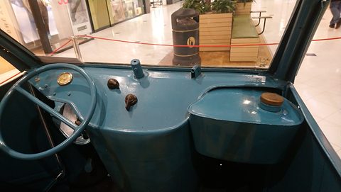 Teal painted dashboard with a large steering wheel, round gauge, two knobs and a capped fuel tank, a shopping-centre floor ahead.