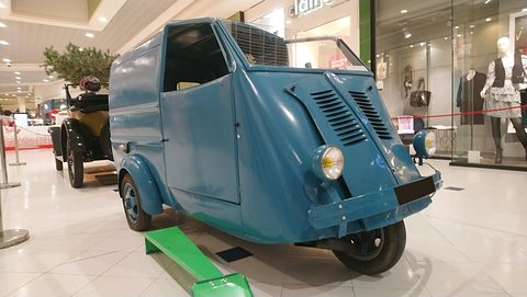 Teal-blue three-wheeled van with louvred nose and twin lamps on a tiled mall floor, a veteran car and shop windows behind. Opens a larger view