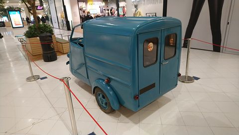 Back of a teal-blue three-wheeled van with two small windowed rear doors, roped off on a shopping-centre floor. Opens a larger view