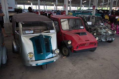 Silver three-wheeled van with a teal louvred nose and a brown canvas back, beside a red boxy microcar under a shed roof. Opens a larger view