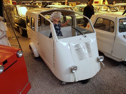White three-wheeled estate van with an open cab, a doll-like mannequin of a child in a bonnet standing inside.