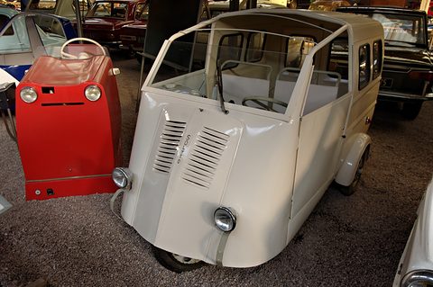 Cream three-wheeled estate van with windows down its side, on gravel next to a small red car. Opens a larger view