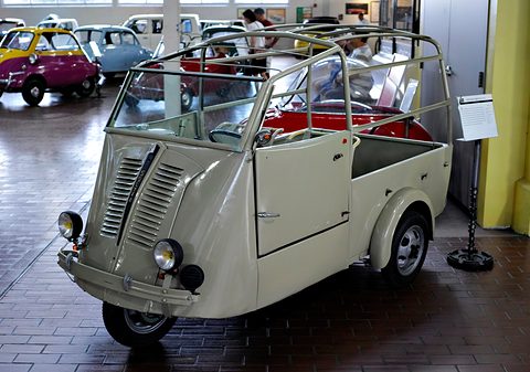 Cream-grey three-wheeled van with a louvred nose and an open tubular roof frame, colourful bubble cars behind it. Opens a larger view