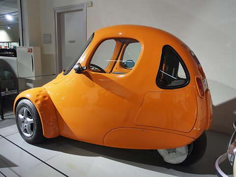Glossy orange teardrop three-wheeler seen from the side and rear, with its single rear wheel, on a pale museum floor. Opens a larger view