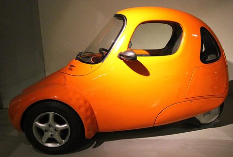 Side view of a glossy orange teardrop-shaped single-seater with a large side window and single rear wheel. Opens a larger view