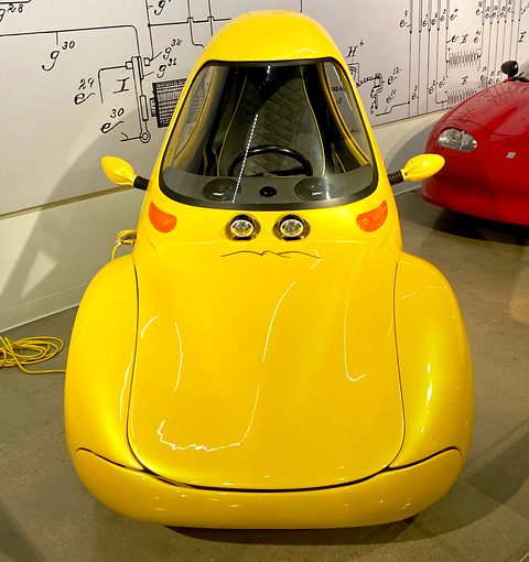 Front view of a yellow-gold teardrop-shaped single-seat car with two small lamps and wing mirrors, patent drawings on the wall. Opens a larger view