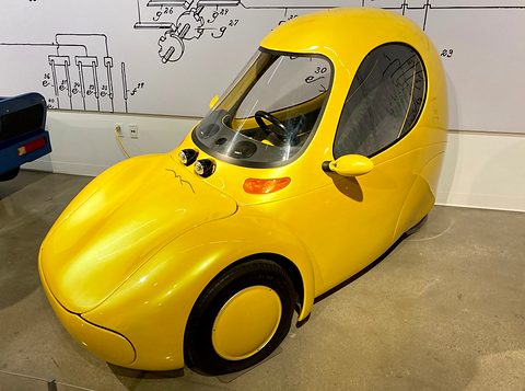A yellow-gold teardrop single-seat car with a large curved windscreen, side door glass and two front wheels. Opens a larger view