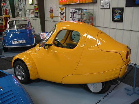 Orange teardrop three-wheeler chained to its stand on a grey floor, a blue bubble car and wall signs behind. Opens a larger view