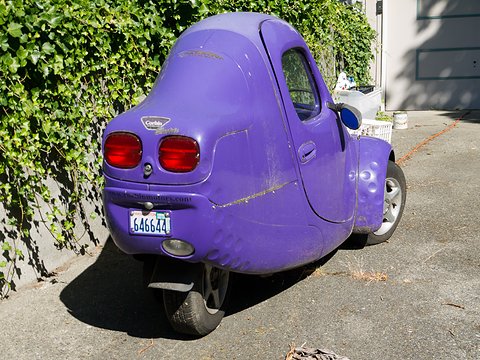 Rear of a worn purple three-wheeler with twin red tail lamps and a single rear wheel casting a shadow. Opens a larger view