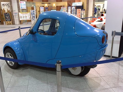 Blue single-seat three-wheeler with a rounded tail and single rear wheel, behind a rope on a marble floor. Opens a larger view
