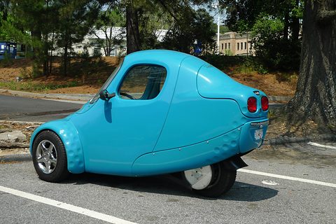 Light-blue single-seat three-wheeler with twin round tail lamps and a single rear wheel on a sunny road. Opens a larger view