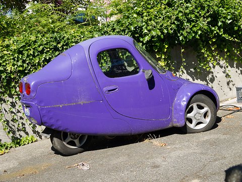 Faded purple single-seat three-wheeler with scuffed paint and alloy front wheel, parked on a driveway beside dense ivy.