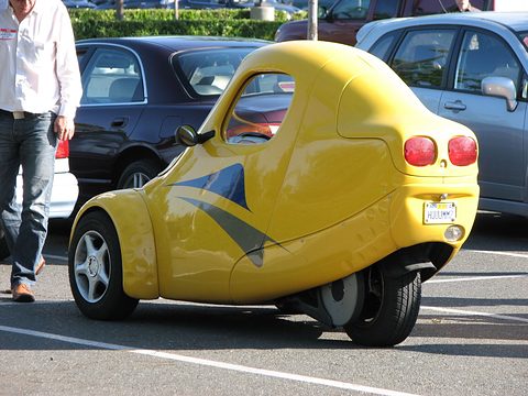 Rear three-quarter of a yellow teardrop-shaped single-seater with twin round tail lamps among parked cars. Opens a larger view